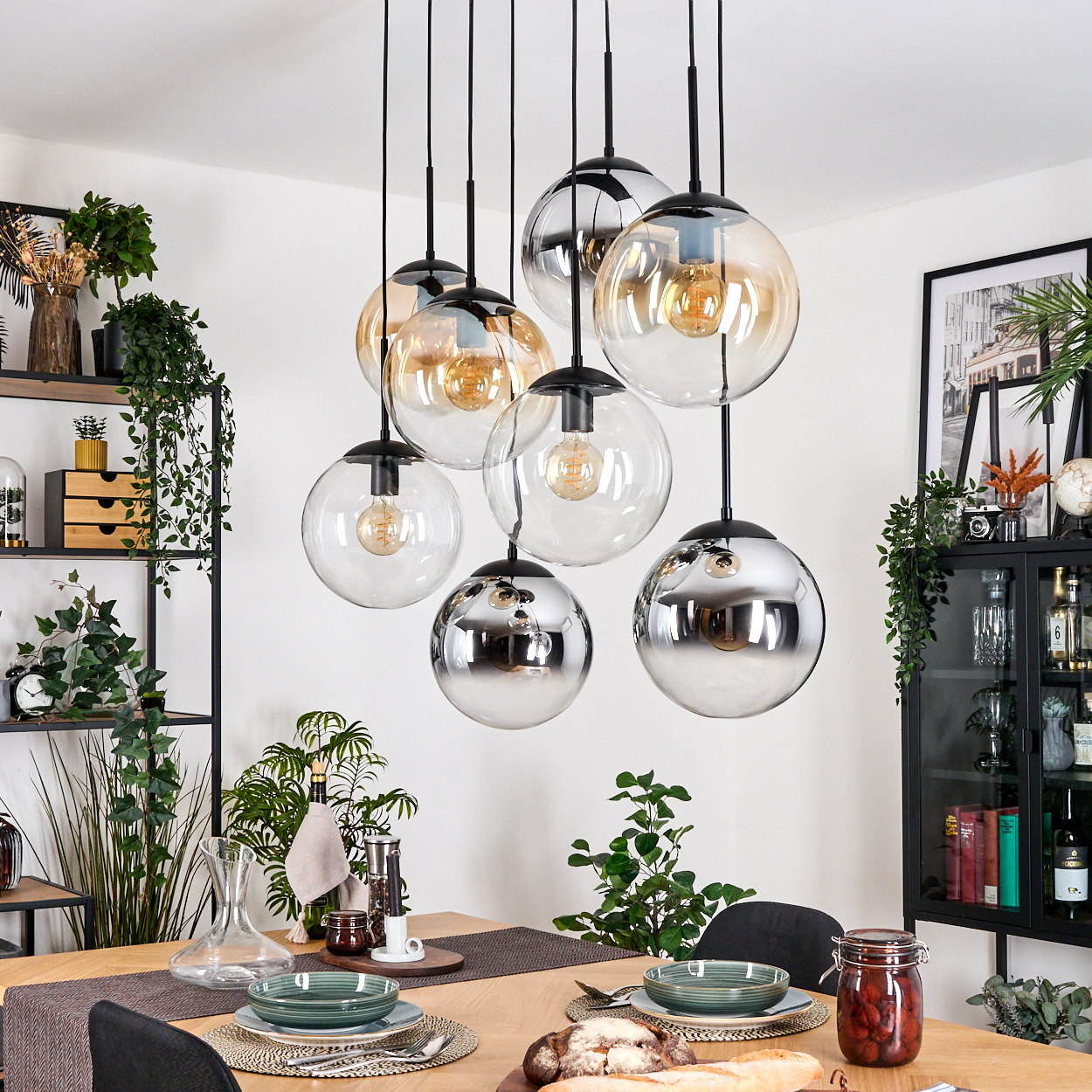 Ein Esszimmer mit Holztisch, Glaskugelleuchten und einem schwarzen Regal mit Pflanzen. Auf dem Tisch stehen Teller und Essen.