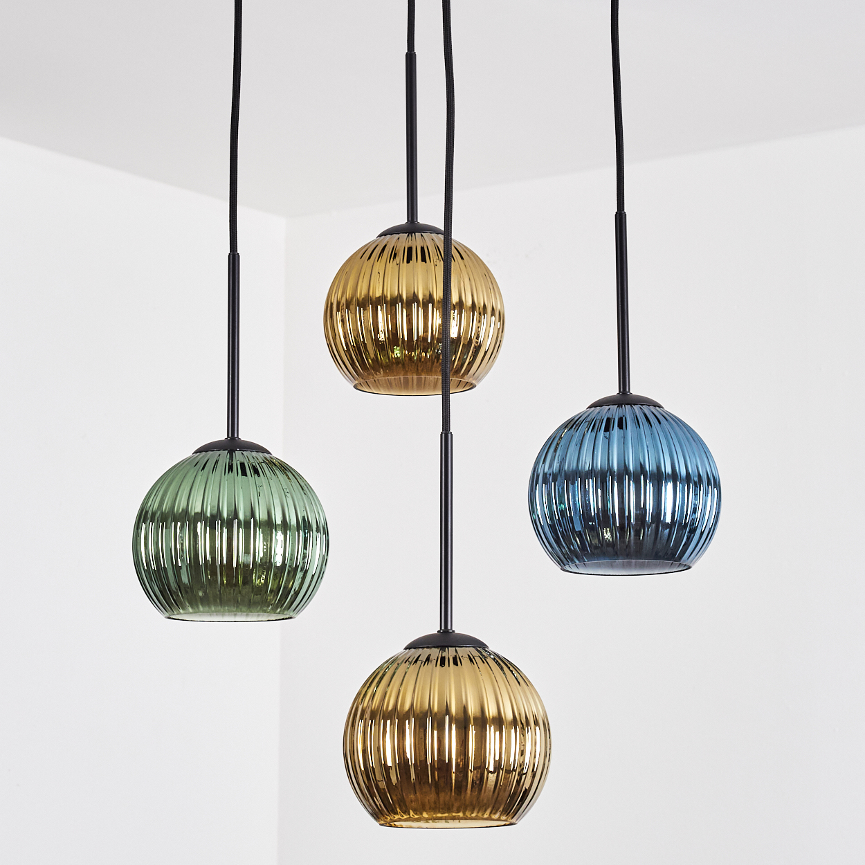 Vier bunte, gerippte Glas-Pendelleuchten hängen vor einer weißen Wand. Farben sind Grün, Gold und Blau. Schwarze Kabel und Armaturen.