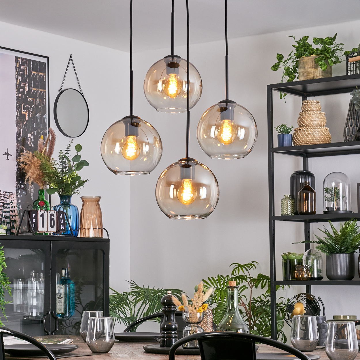 Esszimmer mit Hängeleuchten, Pflanzen und einem schwarzen Metallregal. Tisch mit Gläsern und Dekoration.