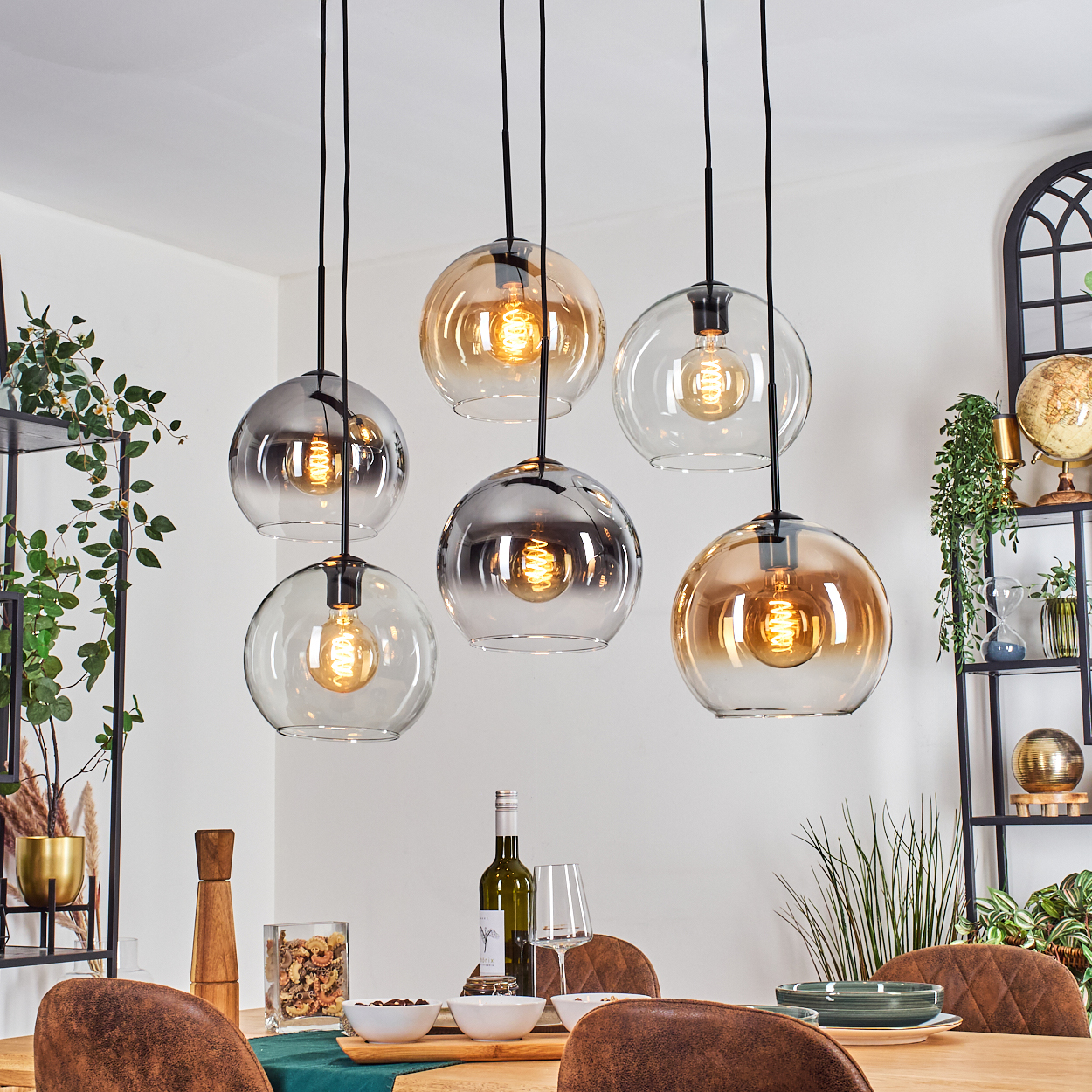 Esszimmer mit sechs hängenden Kugelleuchten, Tisch mit Tellern, Gläsern und Wein. Pflanzen und dekorative Regale sind im Hintergrund.