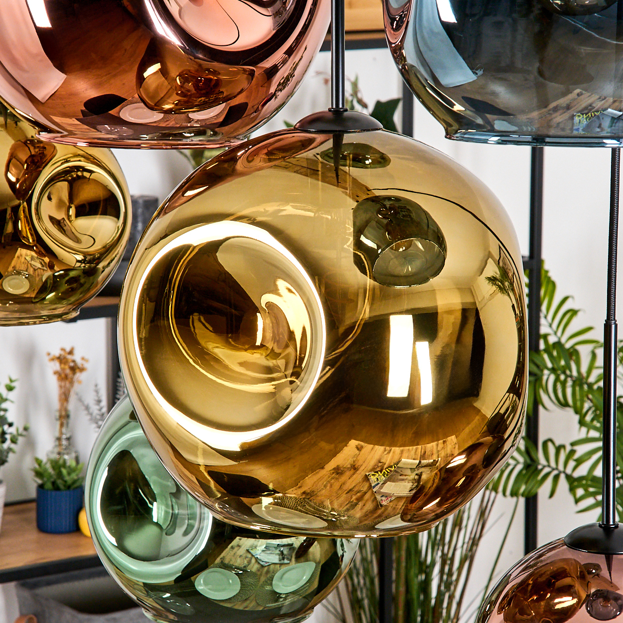 Nahaufnahme von hängenden, kugelförmigen, reflektierenden Pendelleuchten in Gold, Kupfer und Grün. Der Hintergrund umfasst Pflanzen und ein Holzregal.