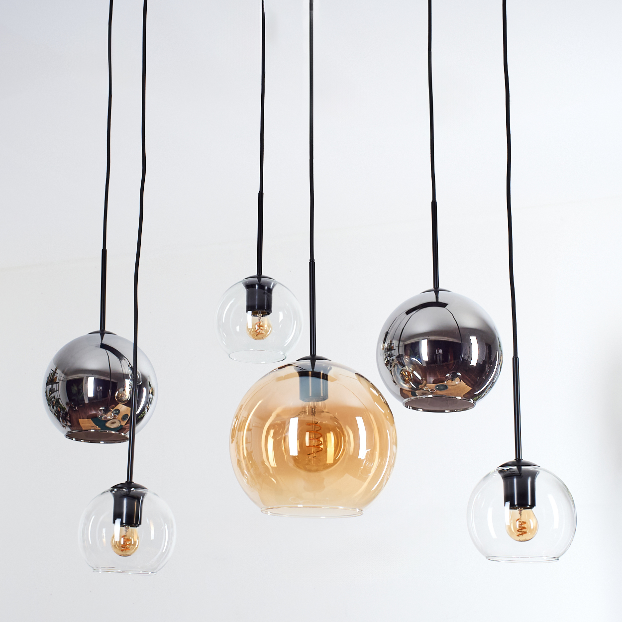Mehrere moderne Pendelleuchten mit Glaskugeln, einige klar, einige verspiegelt und einige bernsteinfarben. Schwarze Kabel hängen von einer weißen Decke.