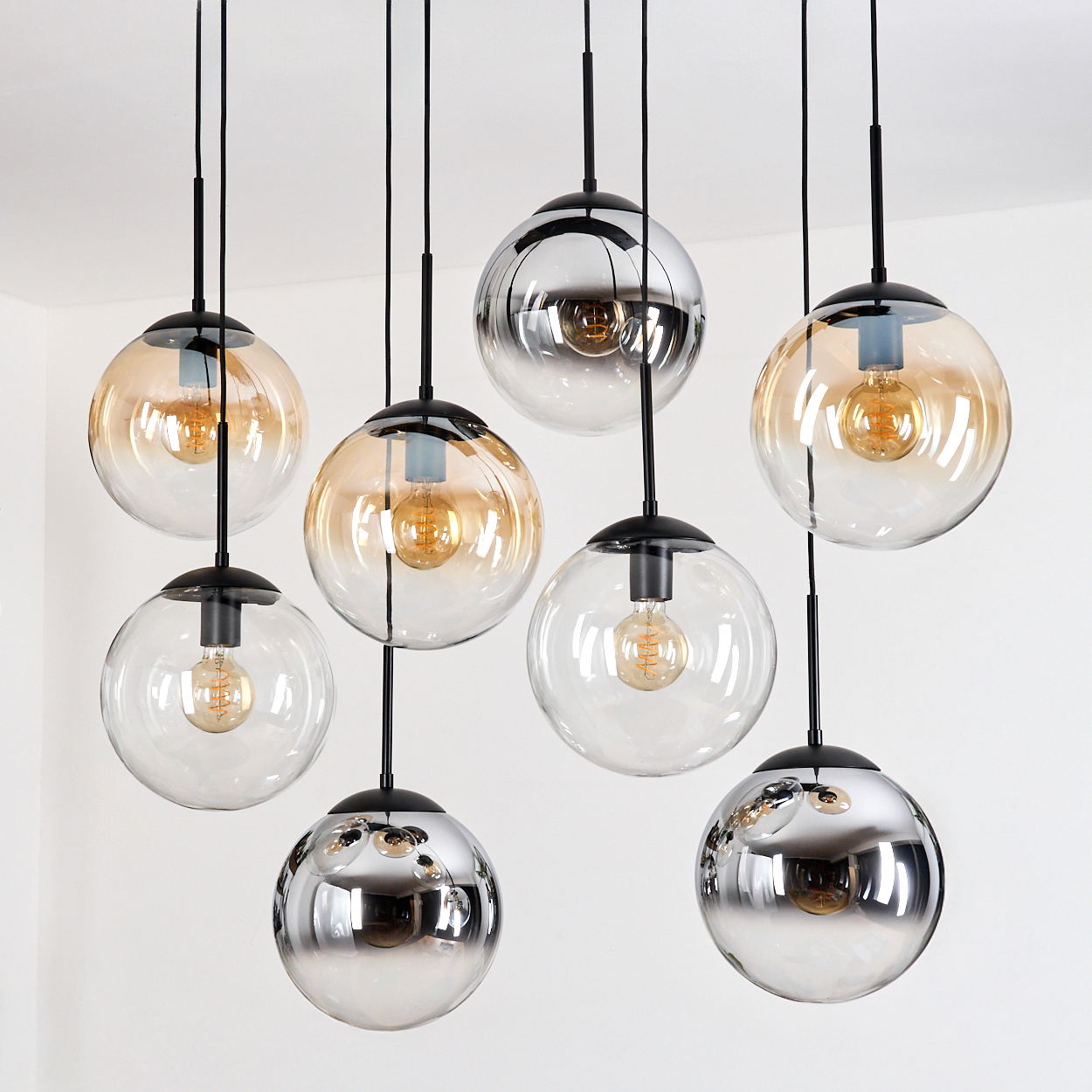 Eine Gruppe kugelförmiger Pendelleuchten mit klaren und bernsteinfarbenen Glasschirmen, schwarzer Hardware und sichtbaren Glühbirnen vor einer weißen Wand.