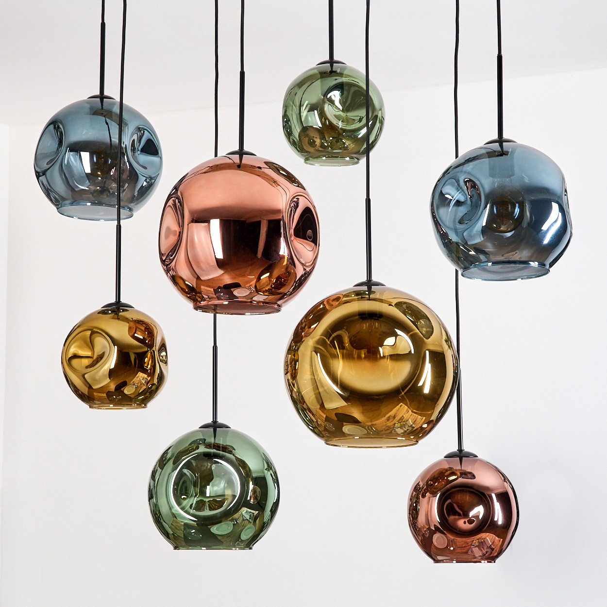 Neun farbenfrohe, kugelförmige Pendelleuchten hängen an schwarzen Schnüren vor einem weißen Hintergrund. Farben sind Blau, Gold und Kupfer.