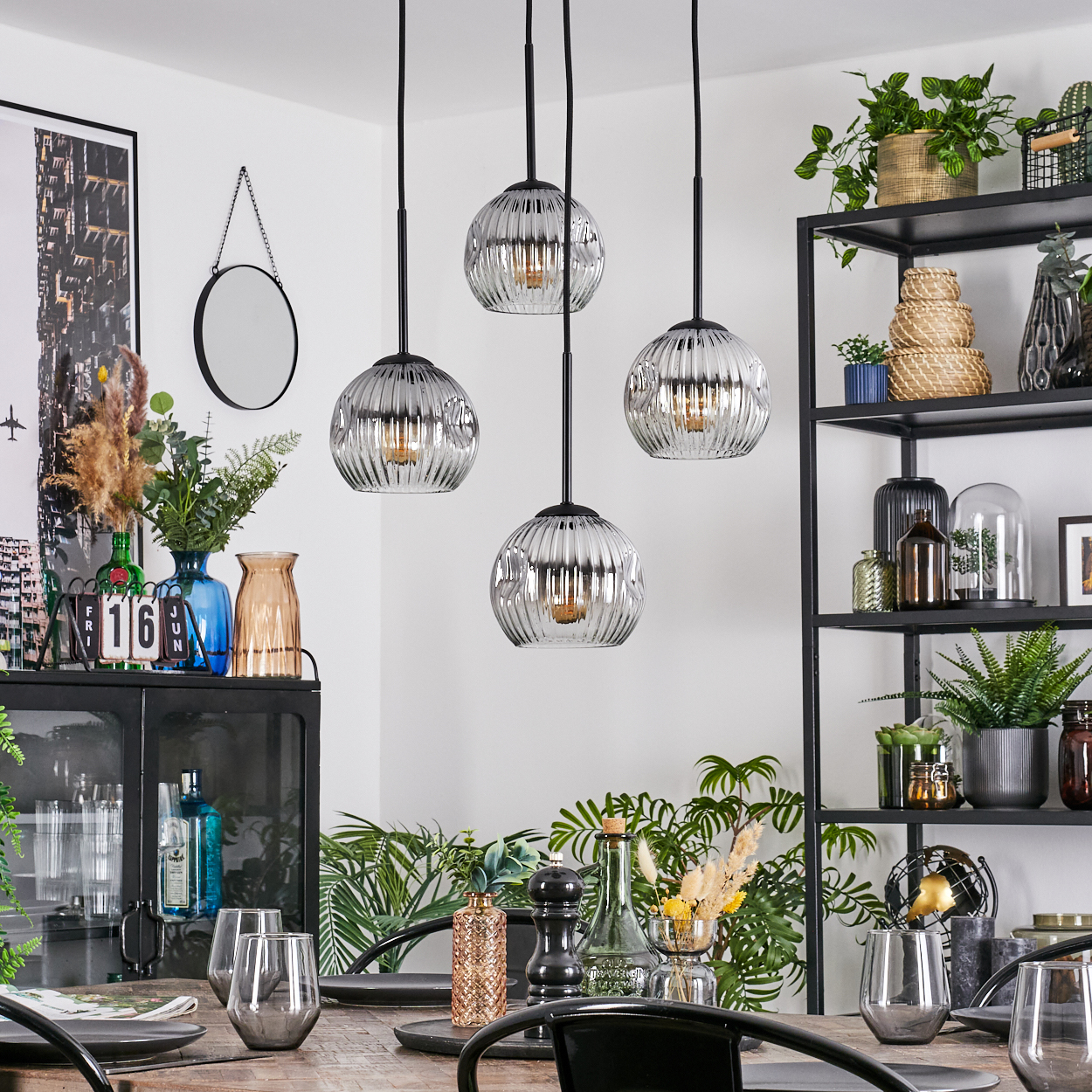Esszimmer mit Hängelampen, Pflanzen und Esstisch. An der Wand befinden sich ein Spiegel und Regale. Gläser stehen auf dem Tisch.