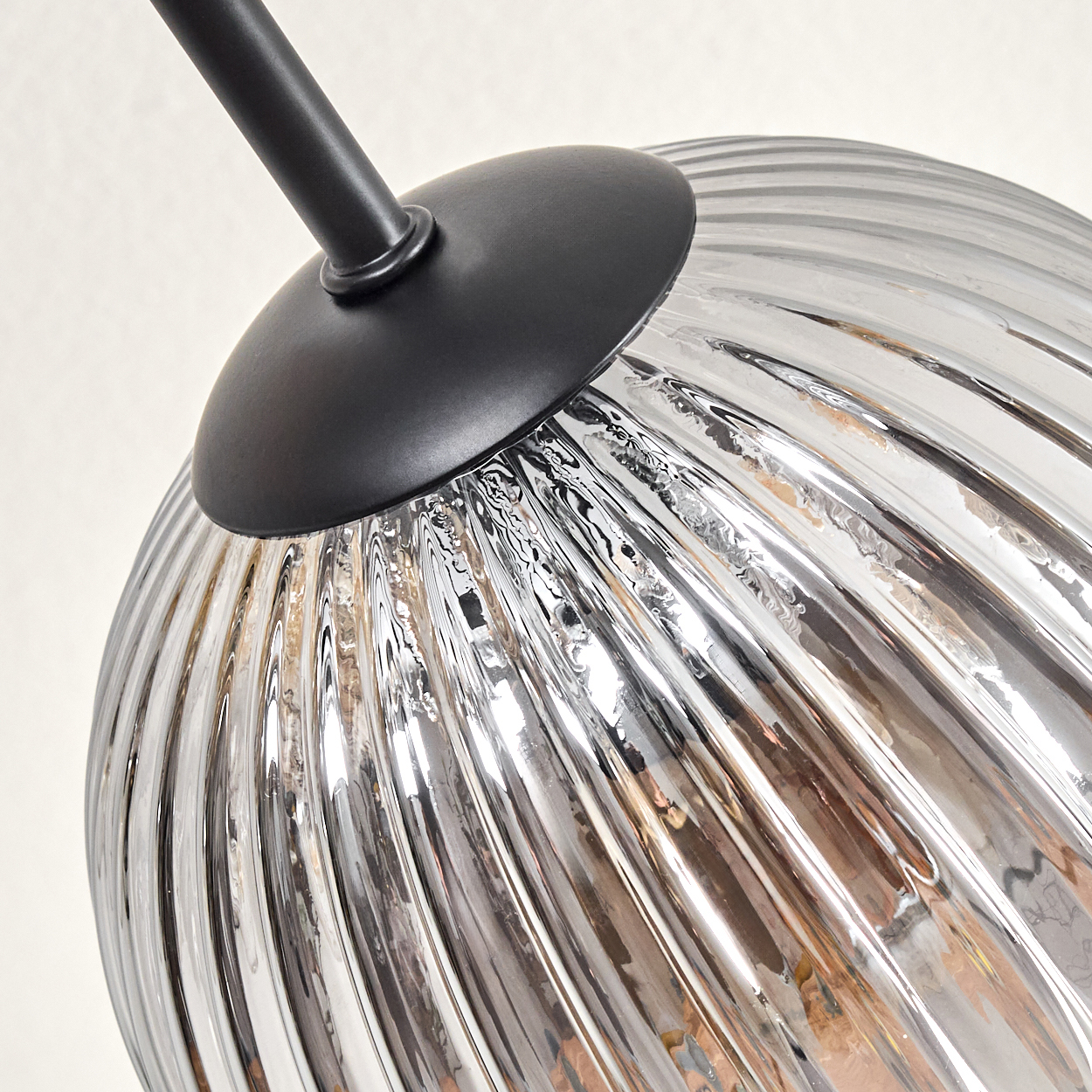Nahaufnahme einer Glasleuchte. Sie hat einen geriffelten, kugelförmigen Glasschirm und eine schwarze Metallbefestigung. Der Hintergrund ist weiß.