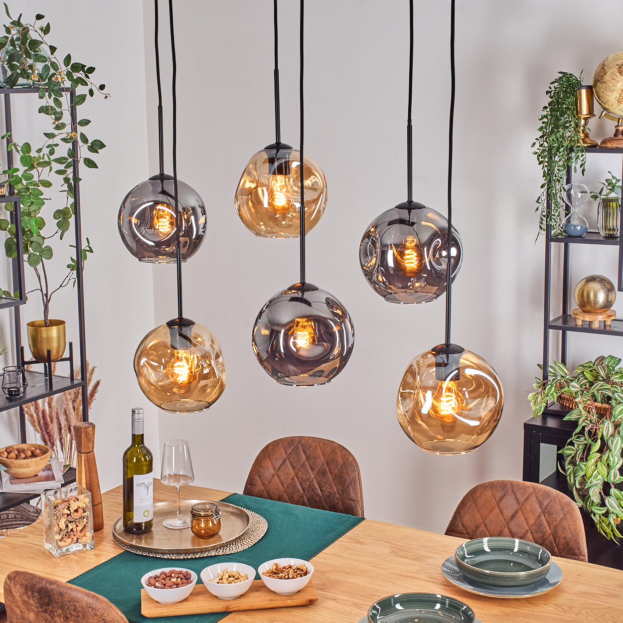 Esszimmer mit sechs hängenden Kugelleuchten. Tisch mit Essen, Wein und Tellern gedeckt. Regale mit Pflanzen und Dekor im Hintergrund.
