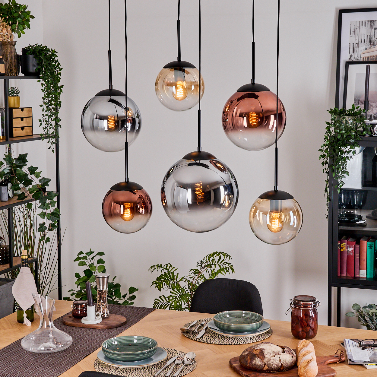 Esszimmer mit Hängeleuchten, Tisch mit Tellern, Brot und Pflanzen. Schwarze Regale mit Büchern und Dekoration.
