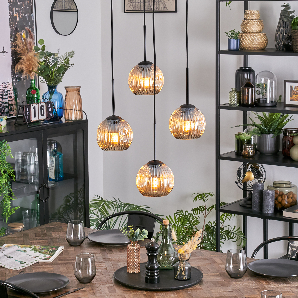 Esszimmer mit Holztisch. Hängelampen, Tischgedeck, schwarzes Regal mit Pflanzen, Vasen und Dekoration.