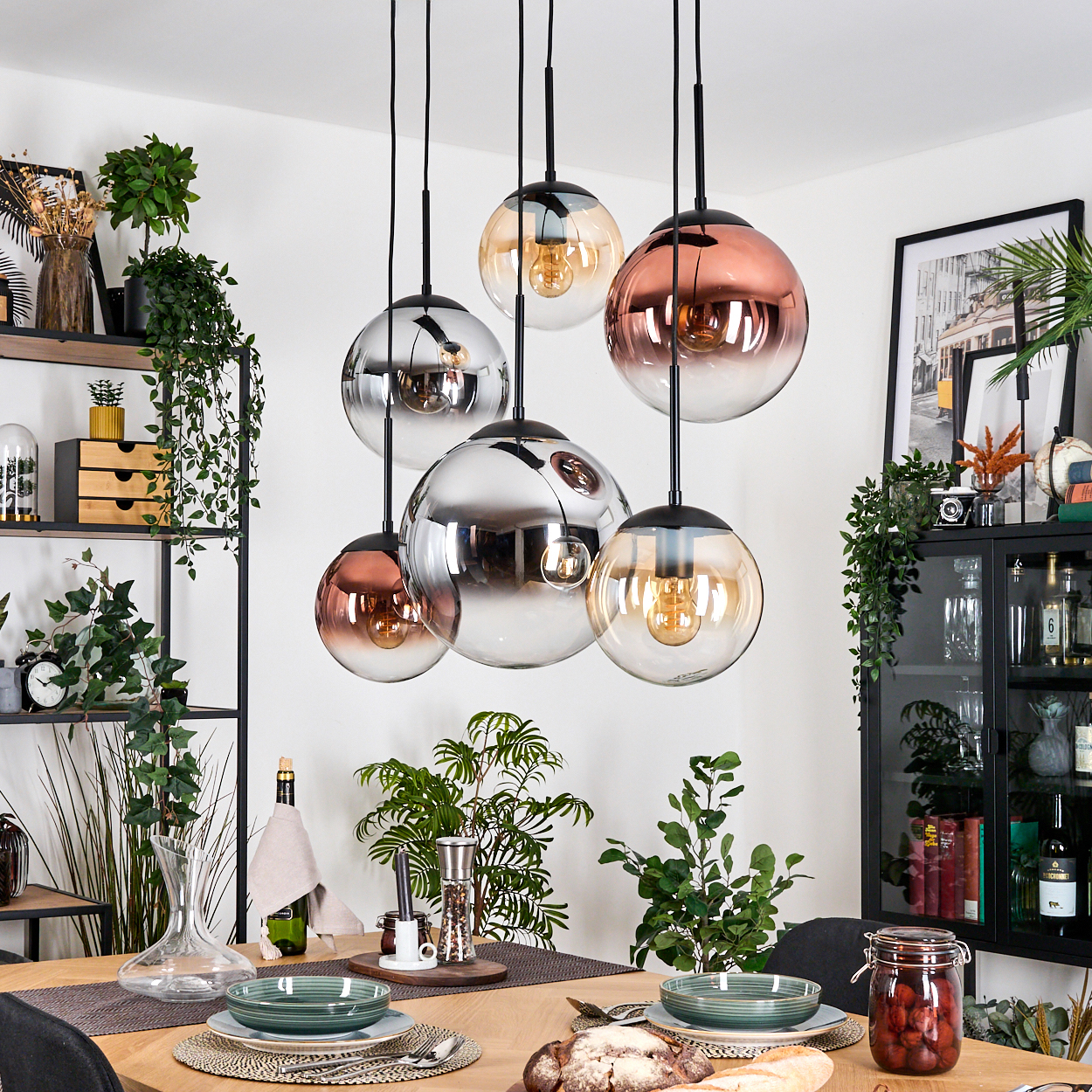 Esszimmer mit Kugel-Pendelleuchten, Tisch mit Tellern und Pflanzen. Im Hintergrund ist ein schwarzes Regal mit Dekor zu sehen.