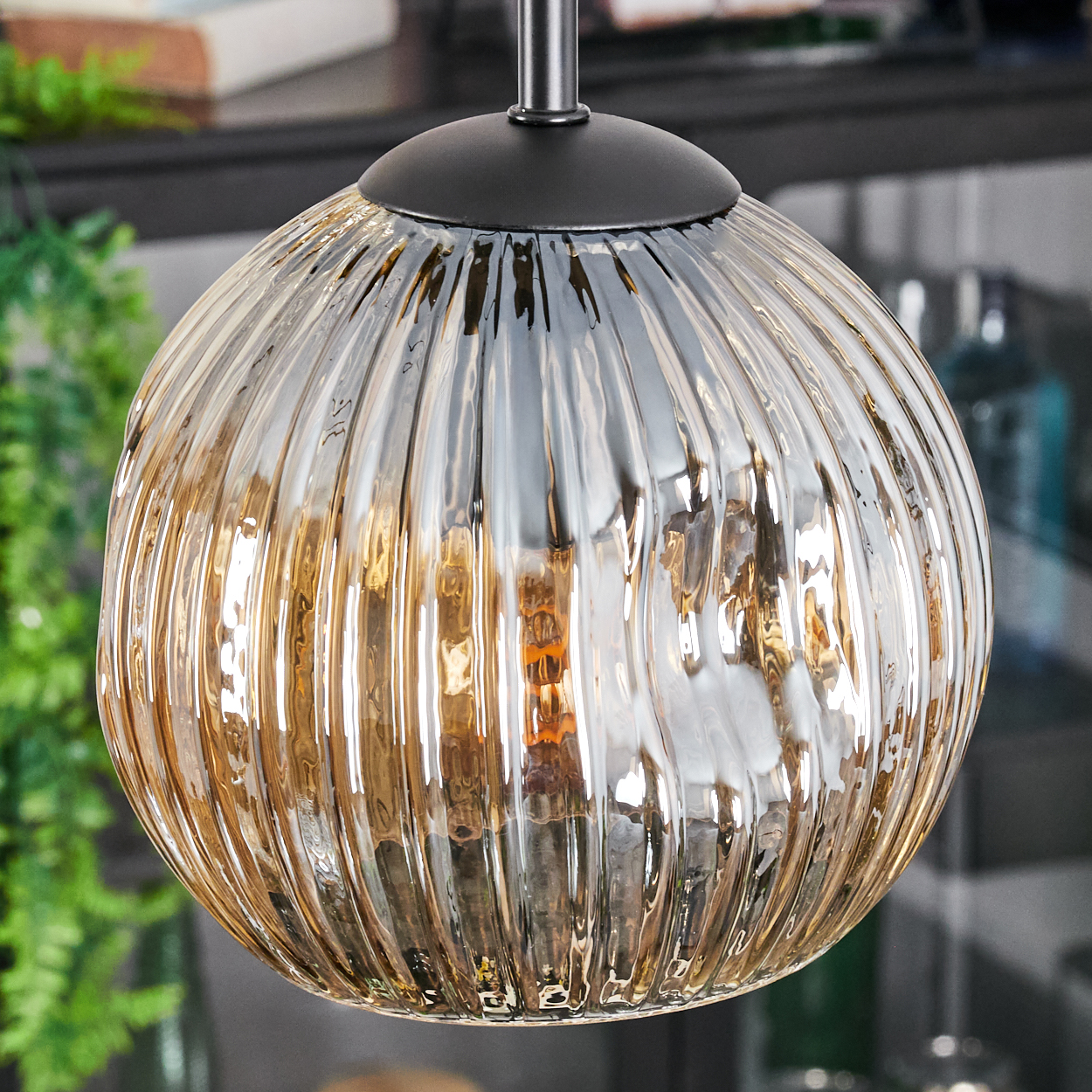 Eine runde, klare Glas-Pendelleuchte mit vertikalen Rippen. Sie hat einen schwarzen Deckel und hängt an einer Metallstange. Der Hintergrund ist unscharf.