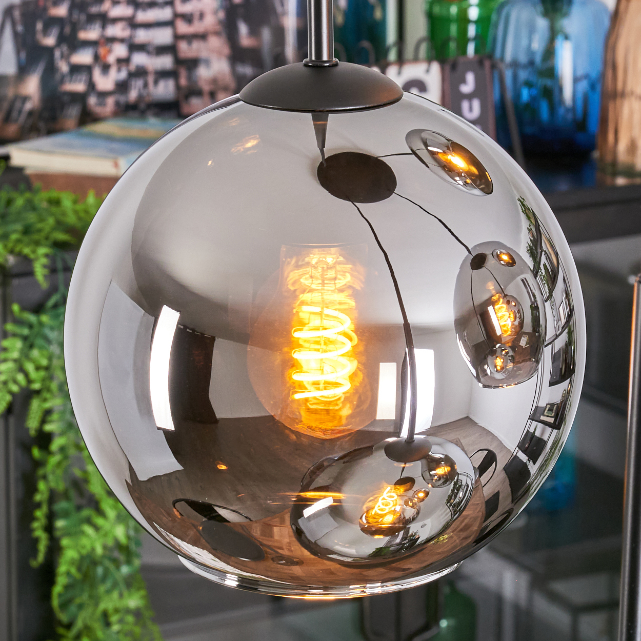 Eine moderne, kugelförmige Pendelleuchte mit reflektierender Oberfläche. Im Inneren leuchtet eine Wendelglühbirne. Der Hintergrund zeigt Dekoration und Pflanzen.