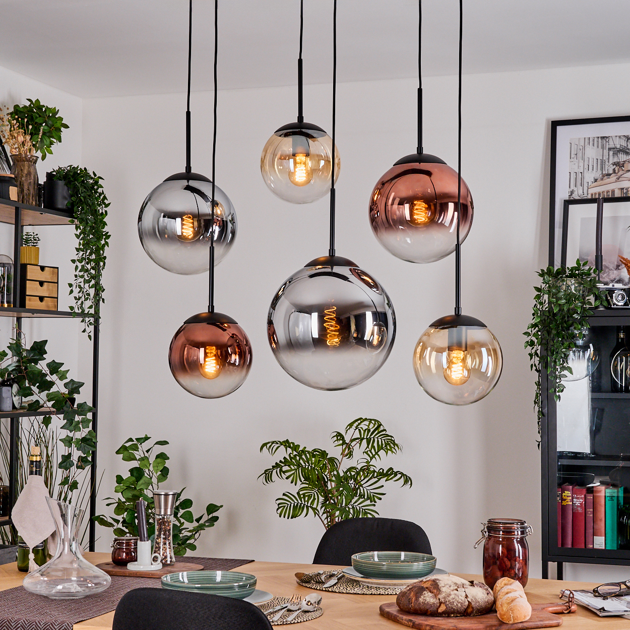 Esszimmer mit sechs hängenden Kugelleuchten. Tisch mit Tellern, Brot und Pflanzen gedeckt. Regale mit Deko und einem gerahmten Bild.