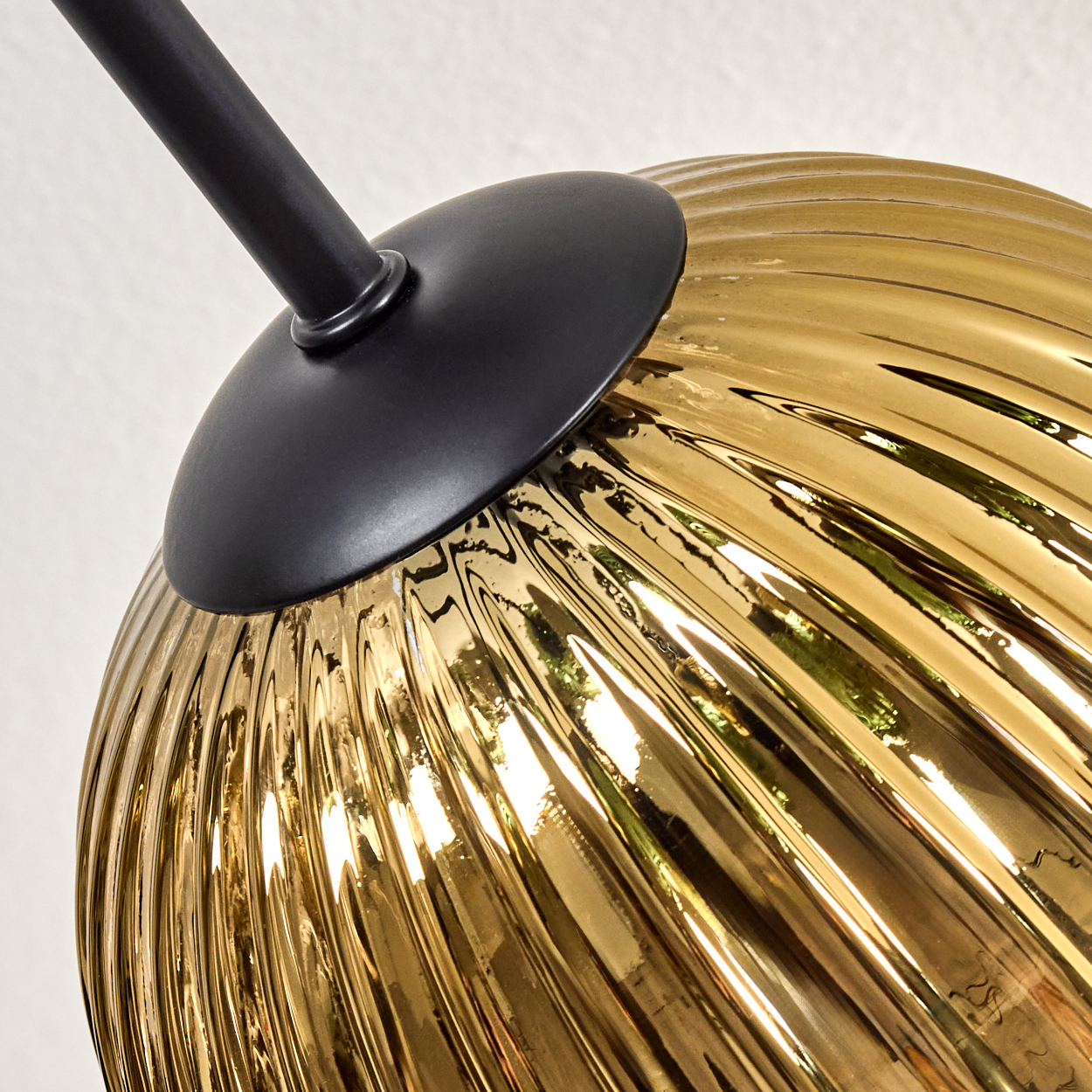 Nahaufnahme eines goldenen Glaslampenschirms mit vertikalen Rillen, schwarzem Metallträger und weißem Hintergrund.