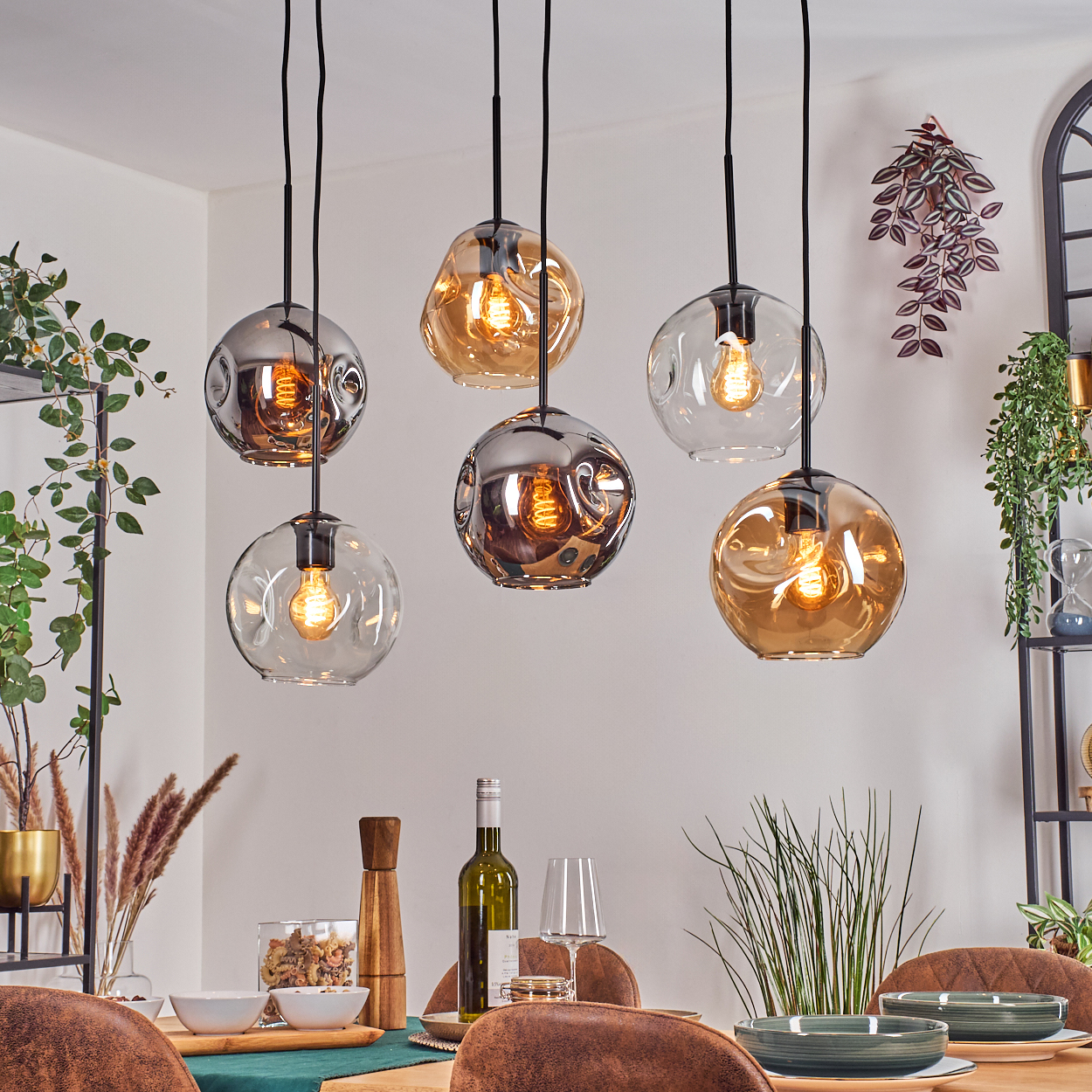 Esszimmer mit hängenden Kugelleuchten, Tisch mit Wein, Gläsern und Tellern. Pflanzen und ein Regal sind im Hintergrund.