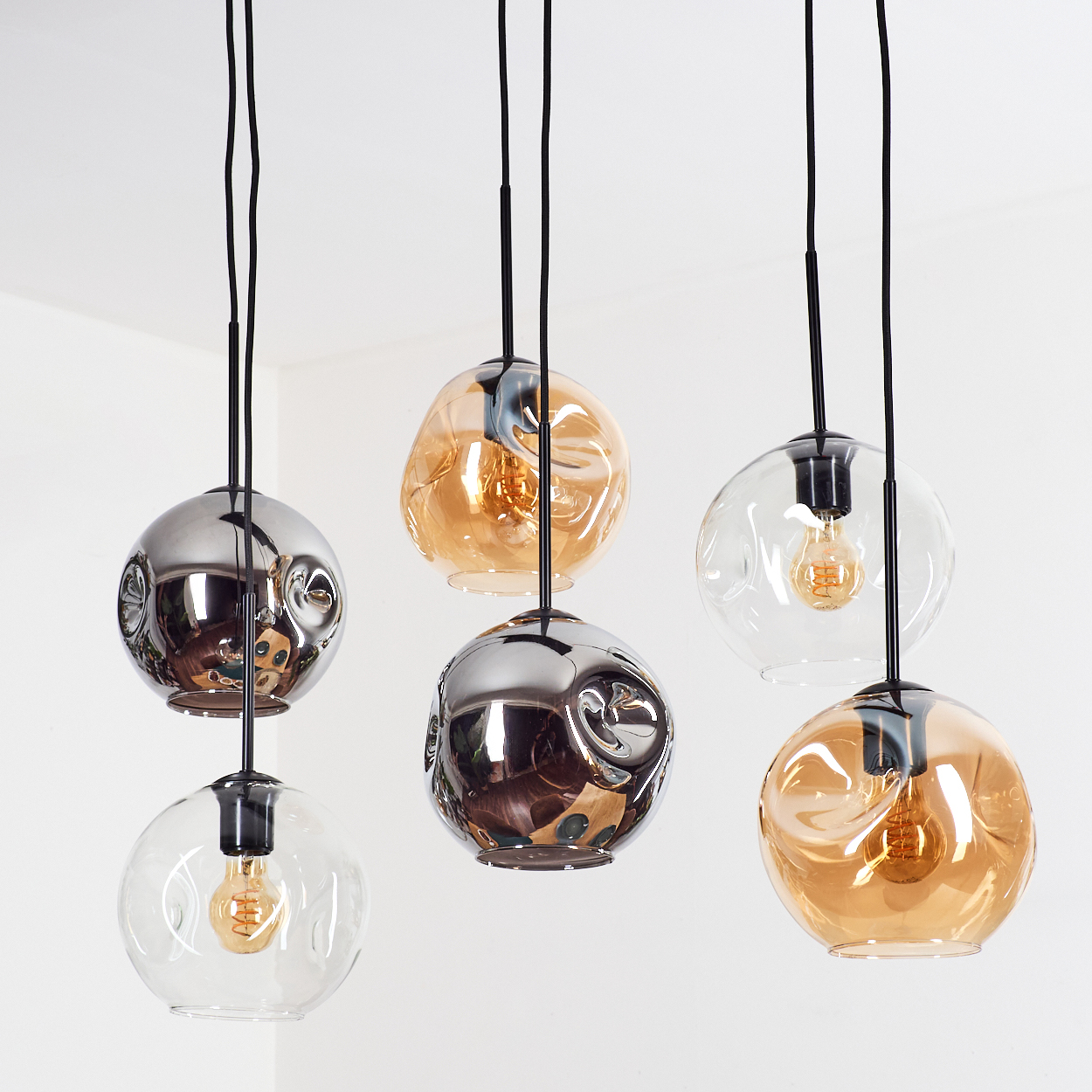 Sechs moderne Pendelleuchten. Einige sind klar, bernsteinfarben oder verchromt. Schwarze Kabel hängen von der Decke vor einer weißen Wand.