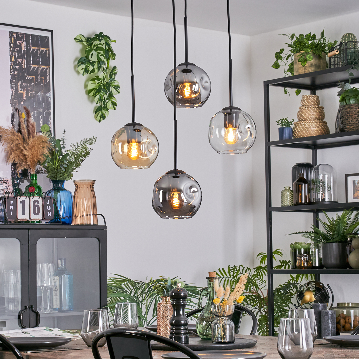 Esszimmer mit Hängelampen, Pflanzen und gedecktem Tisch. Schwarzes Metallregal mit Dekoration. Gläser auf dem Tisch.