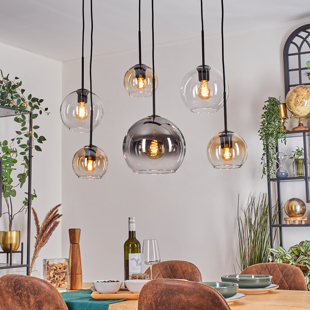 Esszimmer mit hängenden Glaslampen, Tisch mit Tellern, Wein und Stühlen. Regale mit Pflanzen und Dekor im Hintergrund.