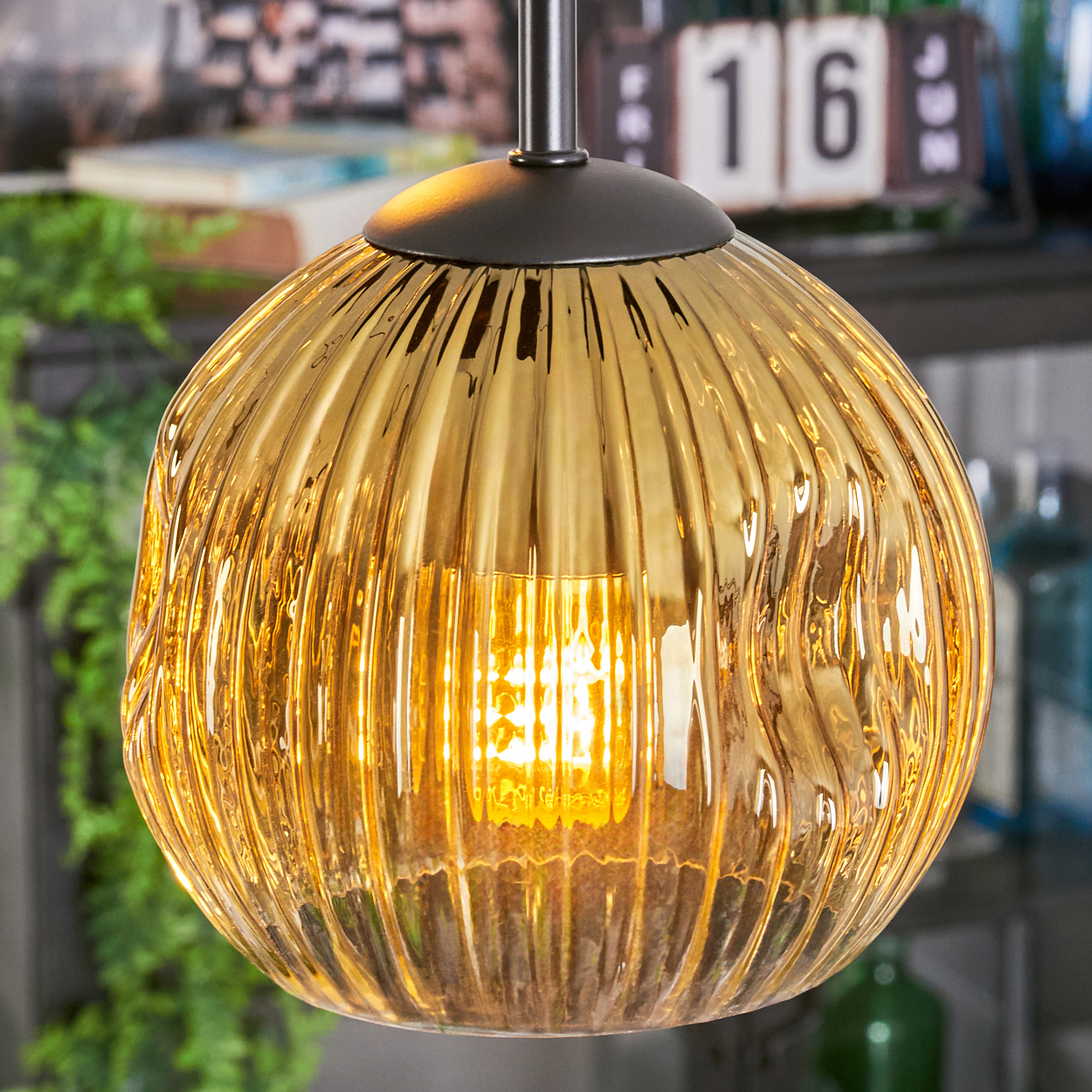 Eine goldene, geriffelte Glas-Hängelampe mit sichtbarer Glühbirne hängt. Der Hintergrund zeigt einen Kalender mit der Zahl 16.