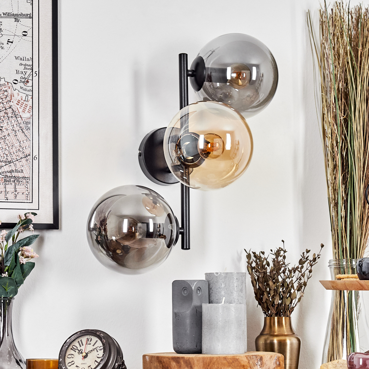 Eine Wandlampe mit drei Glaskugeln. Im Vordergrund befinden sich verschiedene Dekorationen, darunter Kerzen, Vasen und eine Uhr.