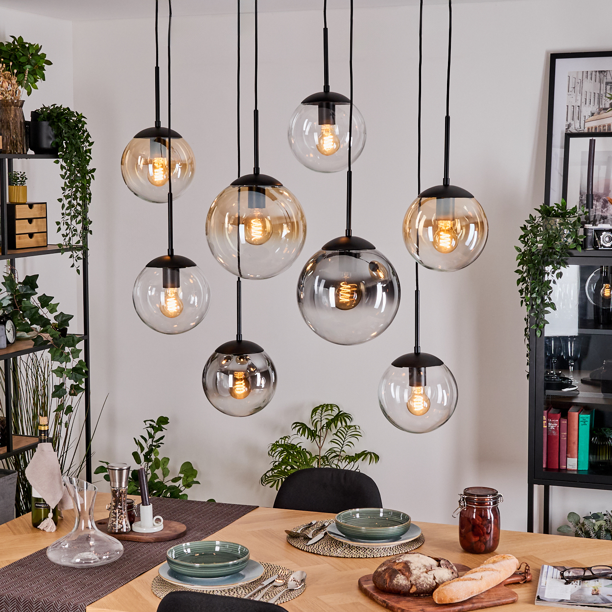 Esszimmer mit hängenden Kugelleuchten. Tisch mit Tellern, Brot und einem Glas. Regale mit Pflanzen und Dekoration sind sichtbar.