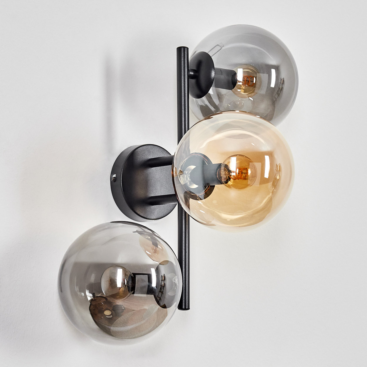 Eine moderne Wandleuchte mit drei kugelförmigen Glasschirmen in Grau und Bernstein, getragen von einem schwarzen Metallrahmen, an einer weißen Wand montiert.