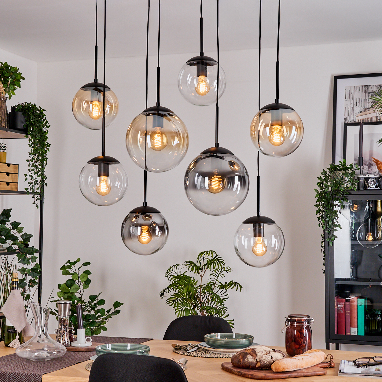 Esszimmer mit hängenden Kugelleuchten. Tisch mit Essen, Pflanzen und Dekoration. Schwarze Regale und ein Schrank sind sichtbar.