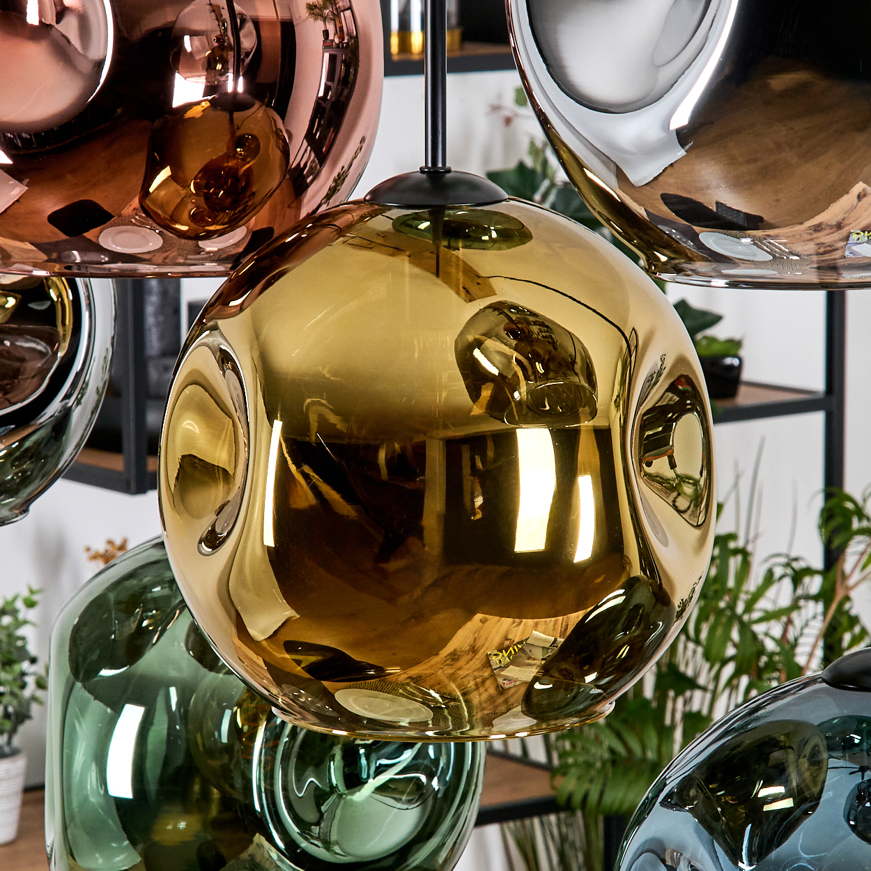 Nahaufnahme von hängenden Glaslampen mit reflektierenden Oberflächen. Farben: Gold, Kupfer, Silber und Grün. Hintergrund: Pflanzen und Regale.