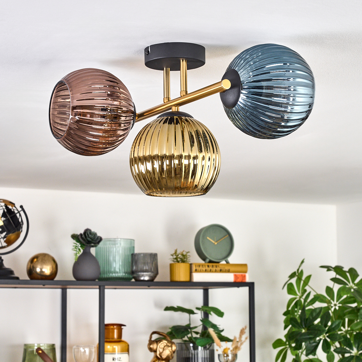 Moderne Deckenlampe mit drei kugelförmigen Schirmen: braun, gold und blau-grau. Regale mit Dekoration sind im Hintergrund sichtbar.