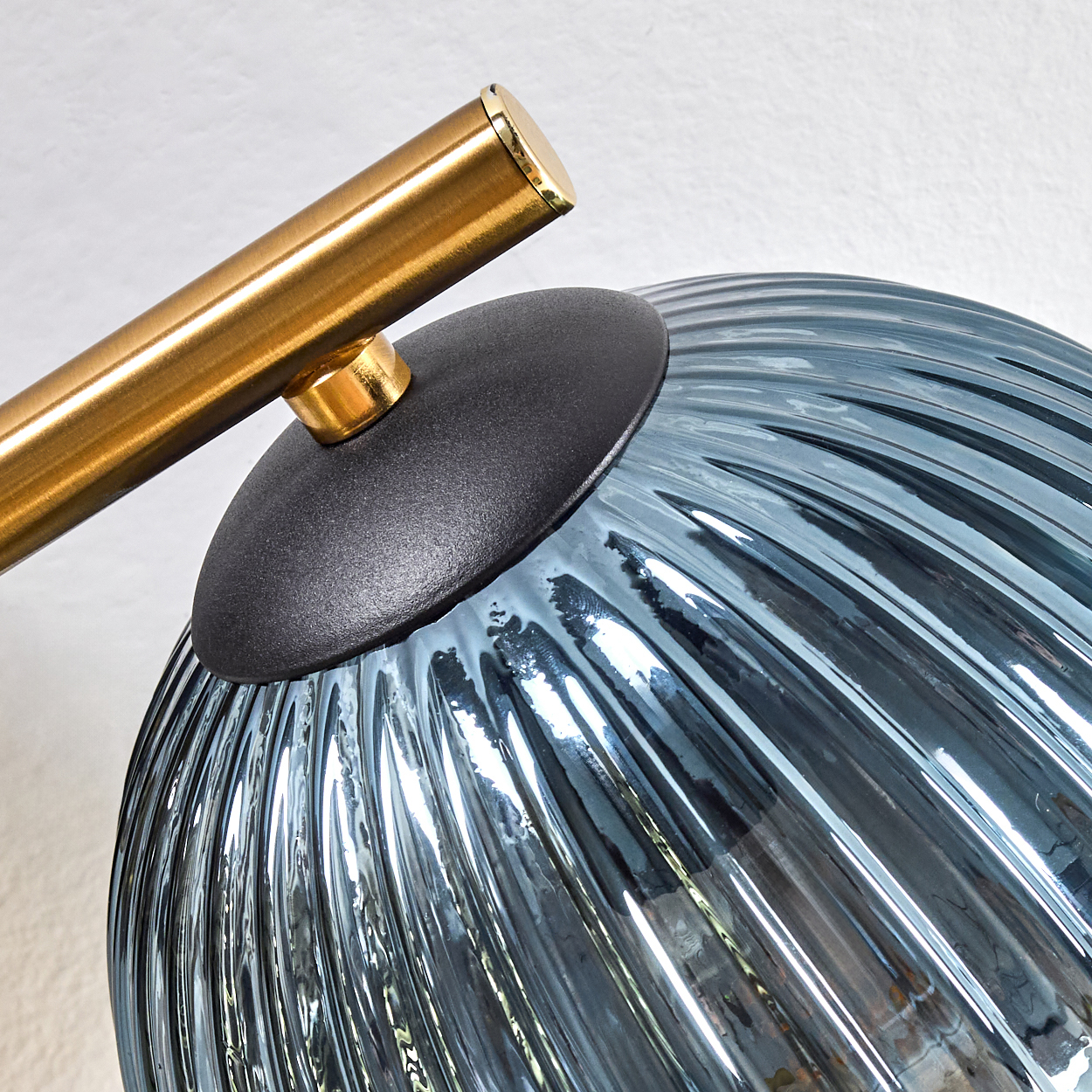 Nahaufnahme einer Lampe. Verfügt über eine Goldstange, eine schwarze Kappe und einen blauen, strukturierten Glasschirm. Der Hintergrund ist eine weiße Wand.