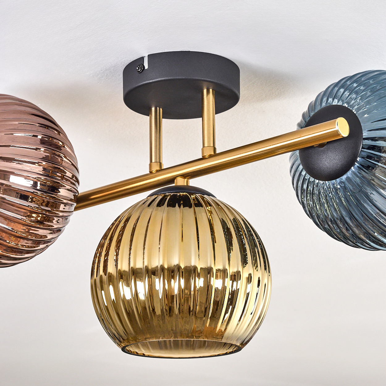 Eine moderne Deckenleuchte mit drei Glaskugeln in Gold, Bronze und Blau, verbunden durch goldene Stäbe, vor weißer Decke.