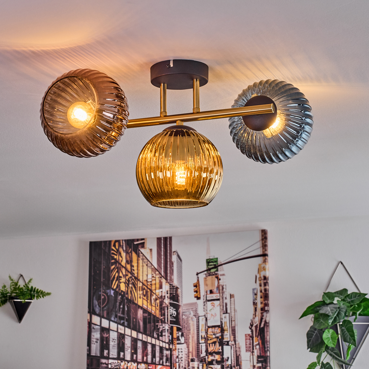 Eine moderne Deckenleuchte mit drei Glasschirmen, golden und grau. Darunter hängt ein Stadtbildgemälde, an den Seiten Pflanzen.