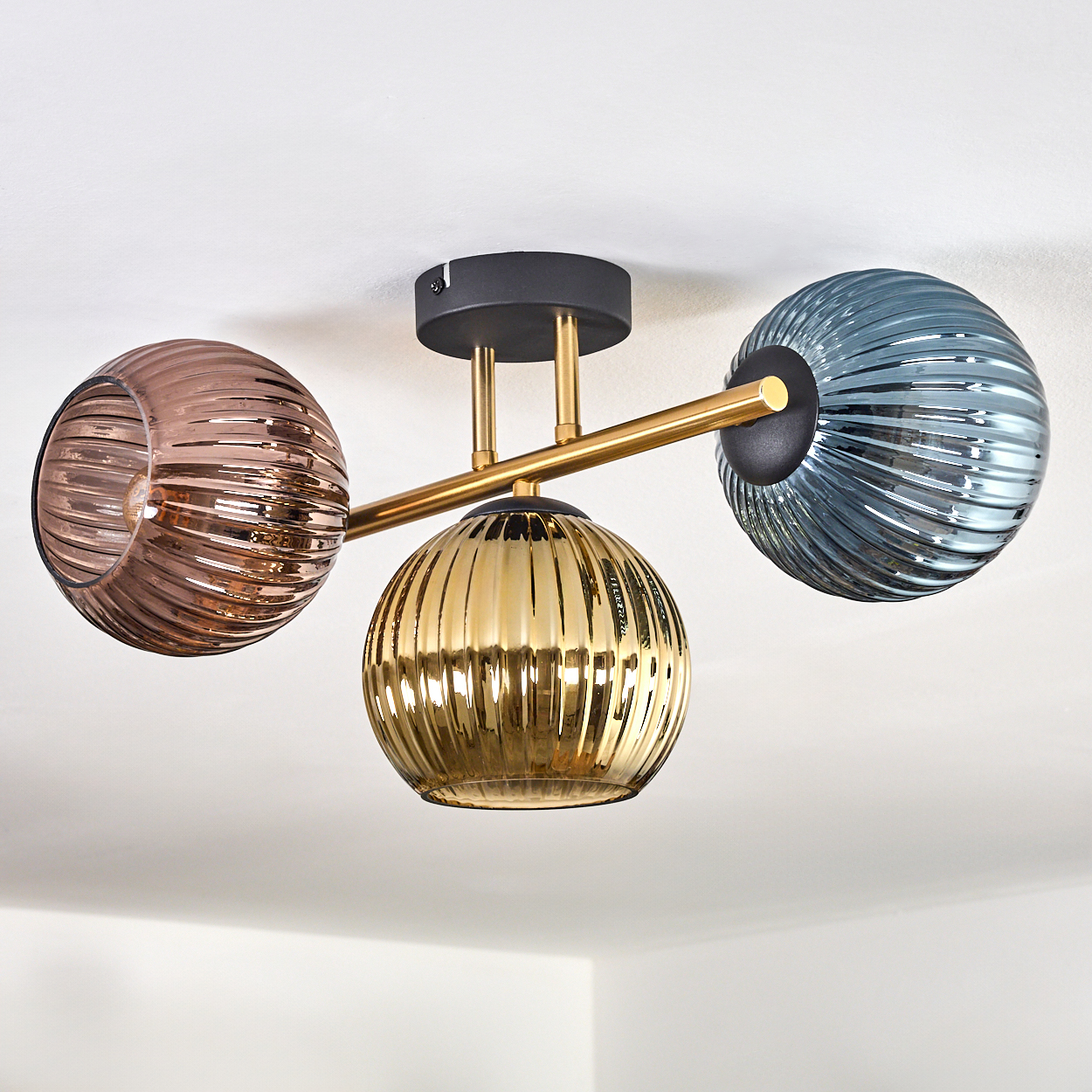 Deckenlampe mit drei gerippten Glaskugeln: braun, gold und blau. Goldfarbene Metallarme und schwarzer Sockel. Weiße Decke.