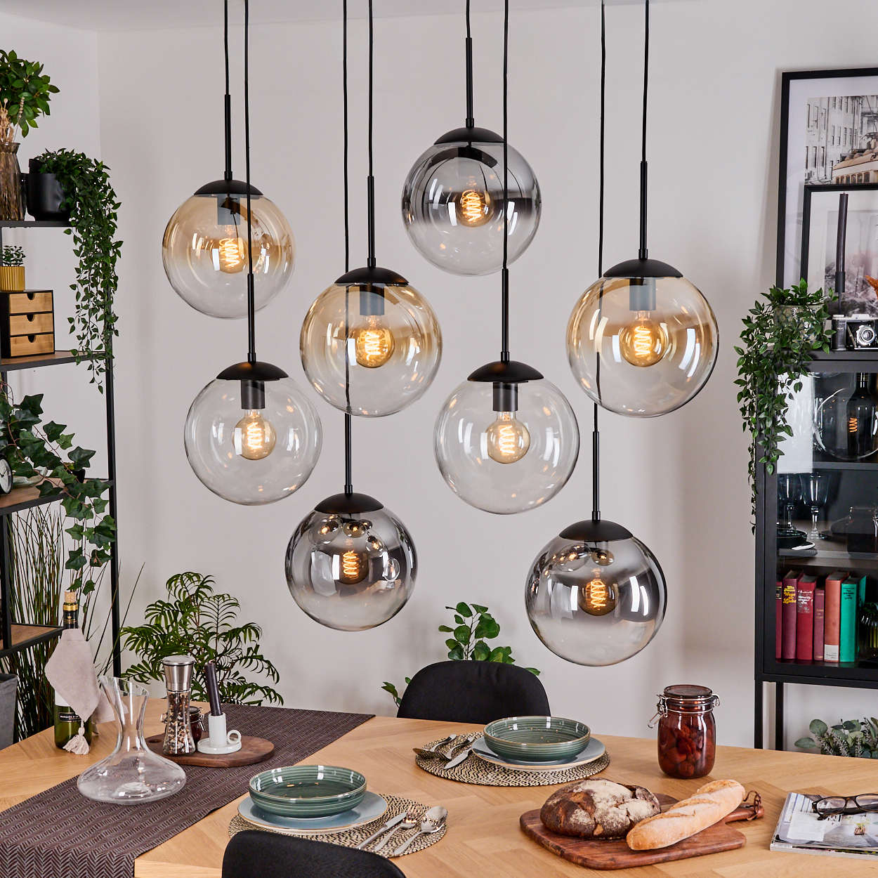 Esszimmer mit hängenden Kugelleuchten. Tisch mit Tellern, Brot und einem Glas. Pflanzen und Regale sind im Hintergrund sichtbar.