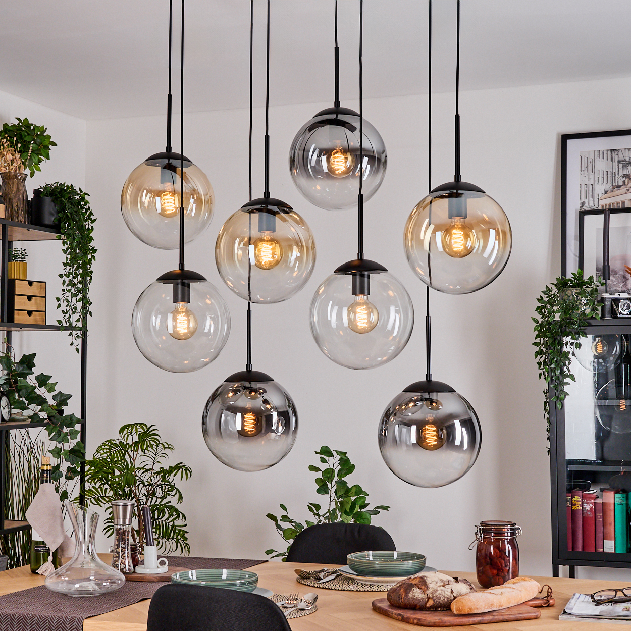 Esszimmer mit acht kugelförmigen Pendelleuchten. Ein Holztisch ist mit Tellern, Essen und Pflanzen gedeckt. Schwarze Regale im Hintergrund.