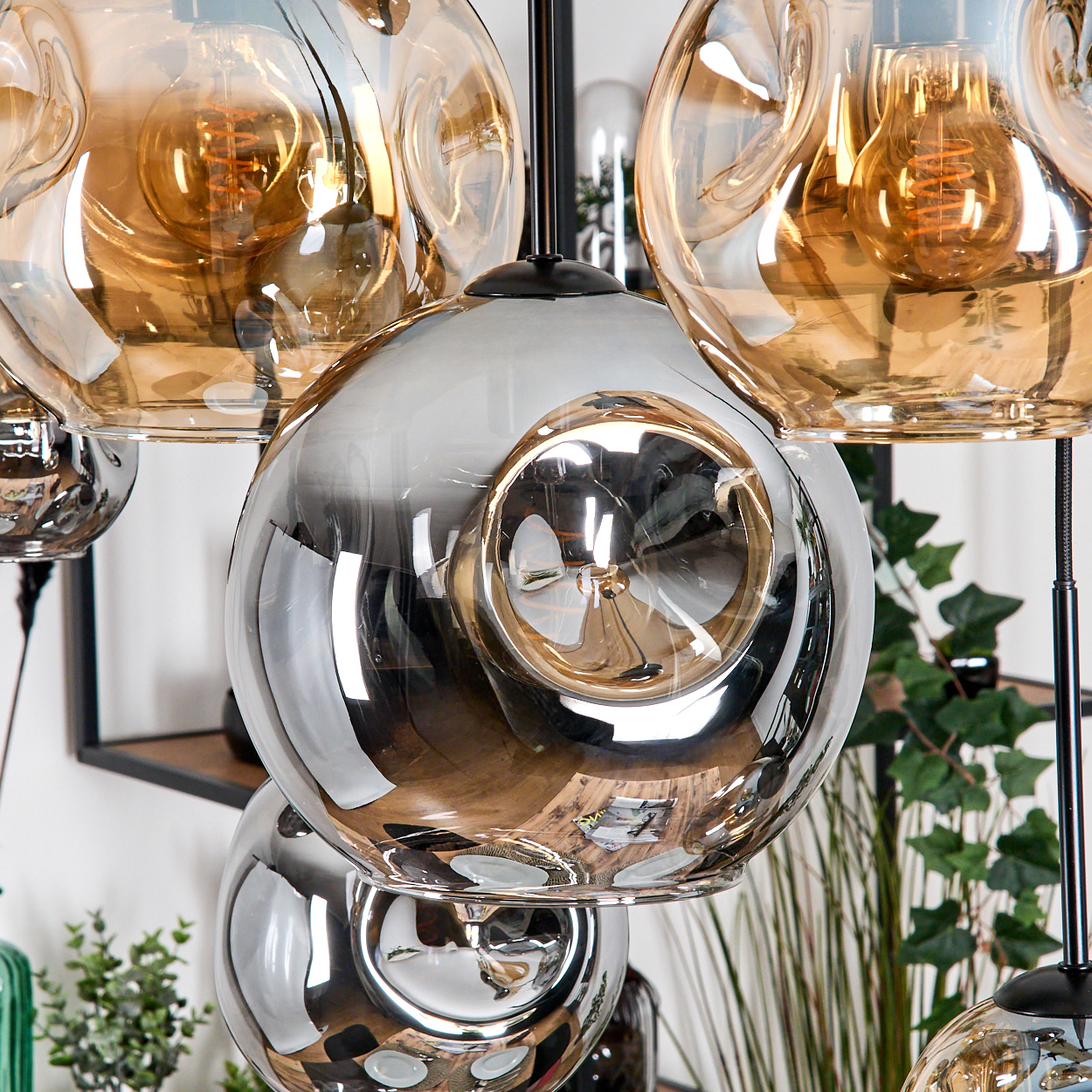 Nahaufnahme von modernen Glas-Pendelleuchten. Glühbirnen innen. Metallische Silber- und Bernsteinfarben. Pflanzen im Hintergrund.