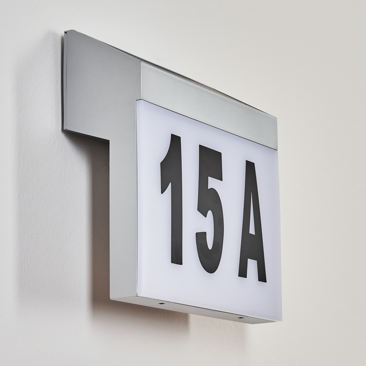 Ein rechteckiges Schild mit der schwarzen Nummer 15A auf weißem Hintergrund, montiert an einer weißen Wand. Das Schild hat einen silbernen Rahmen.