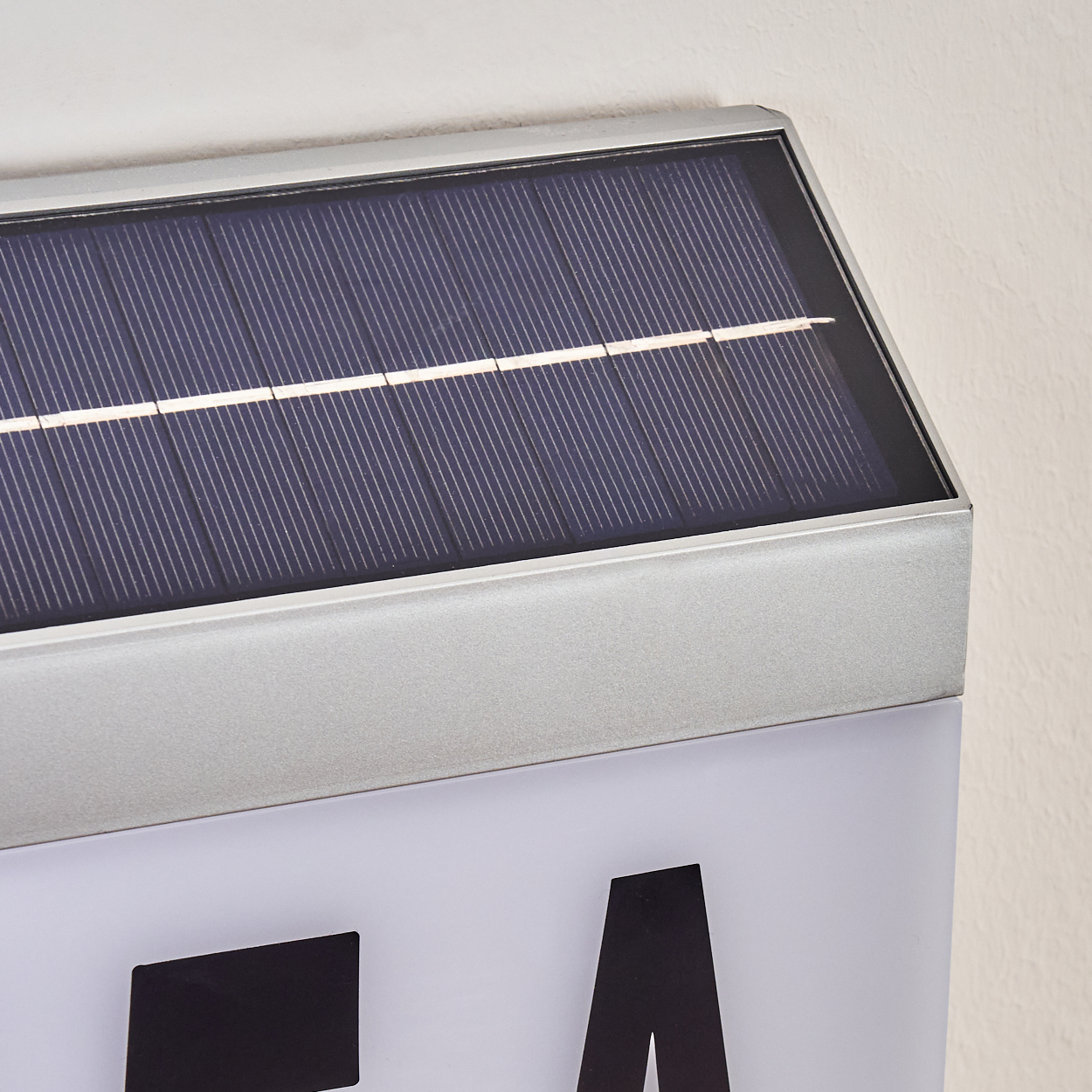 Nahaufnahme eines Solarpanels auf einem Schild. Das Panel ist schwarz und silber, das Schild weiß mit schwarzen Buchstaben. Die Wand ist hell.