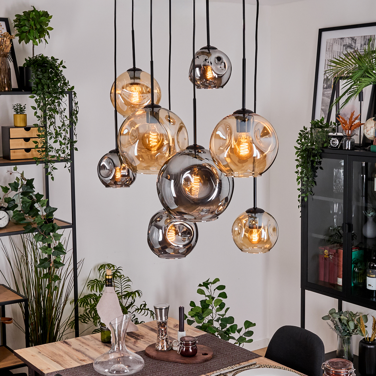 Esszimmer mit Holztisch, hängenden Blasenlichtern, Pflanzen und einem schwarzen Schrank. Auf dem Tisch stehen eine Karaffe und eine Pfeffermühle.
