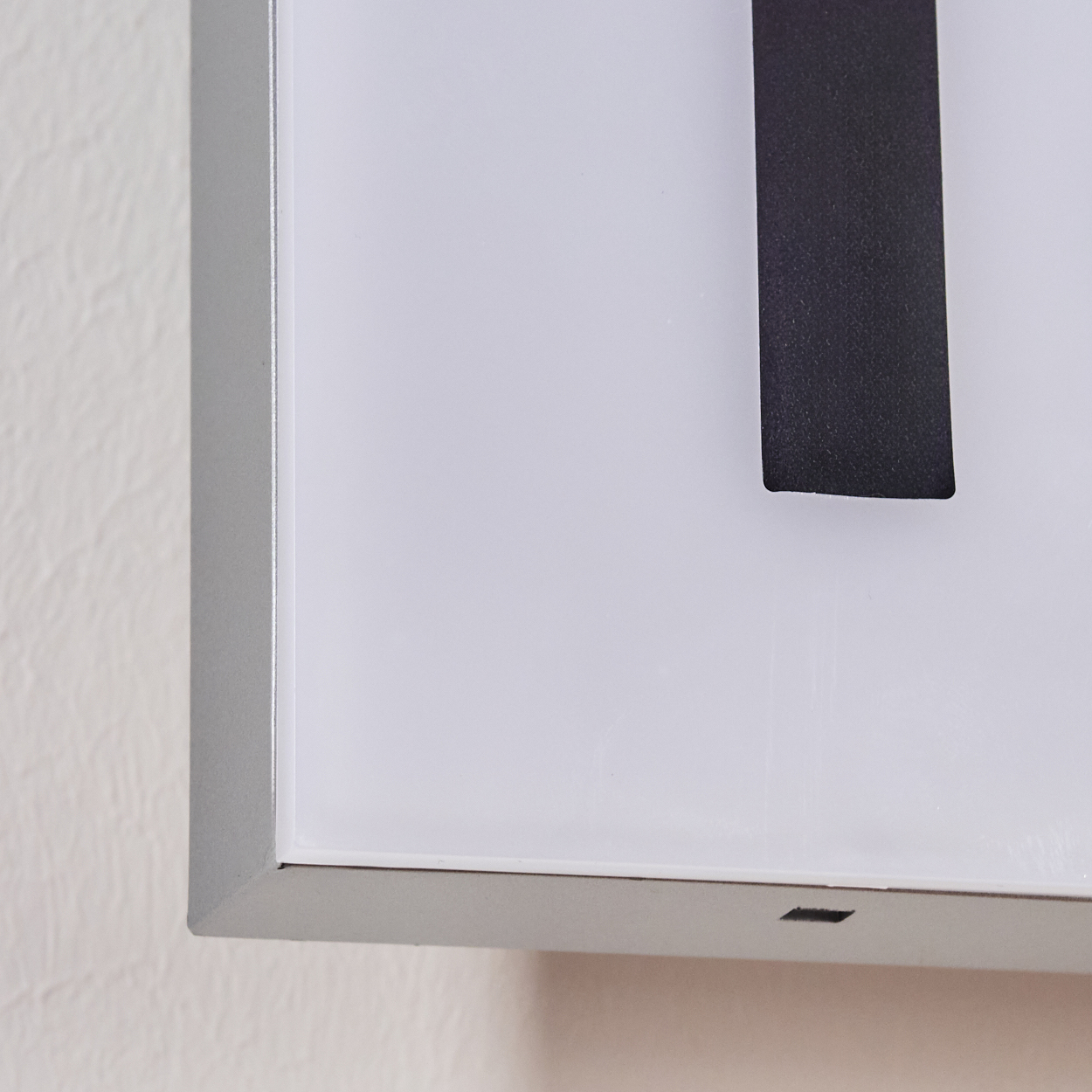 Nahaufnahme eines weißen Zifferblatts mit einem schwarzen Zeiger. Ein silberner Rahmen umgibt die Uhr. Die Wand ist beige und strukturiert.