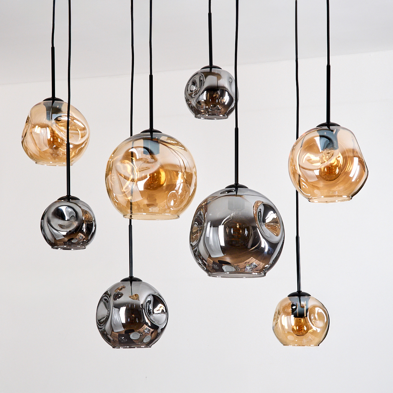 Mehrere Glas-Pendelleuchten hängen von der Decke. Einige sind bernsteinfarben, andere silberfarben. Alle haben schwarze Kabel und sind beleuchtet.
