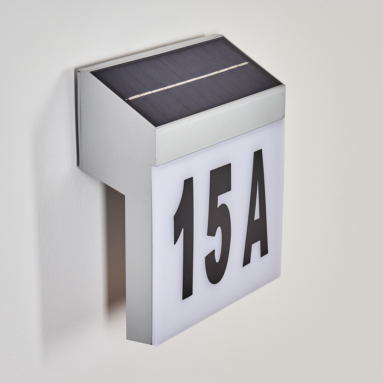 Ein solarbetriebenes Adressschild. Es zeigt "15A" in Schwarz auf weißem Grund. Das Schild ist an einer hellgrauen Wand montiert.