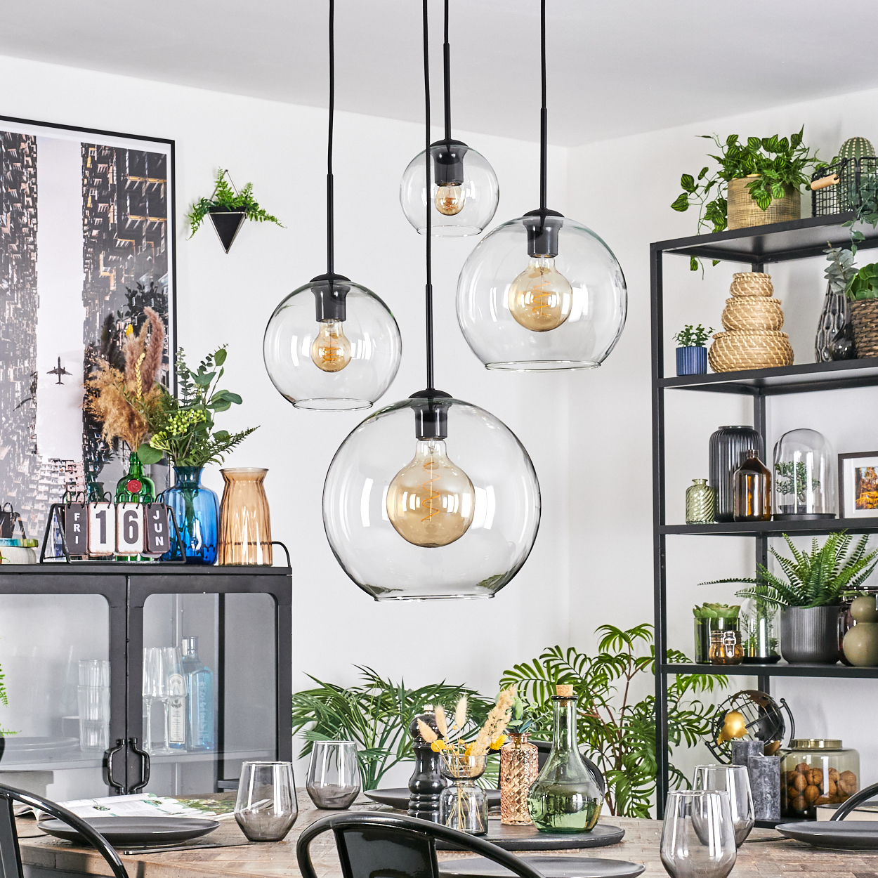 Esszimmer mit Hängekugelleuchten, Tisch mit Gläsern und einem schwarzen Metallregal mit Pflanzen und Dekoration.