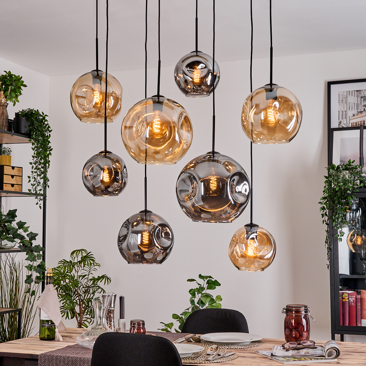 Esszimmer mit hängenden Kugelleuchten. Tisch mit Tellern, Gläsern und Dekorationen. Pflanzen und Regale im Hintergrund.