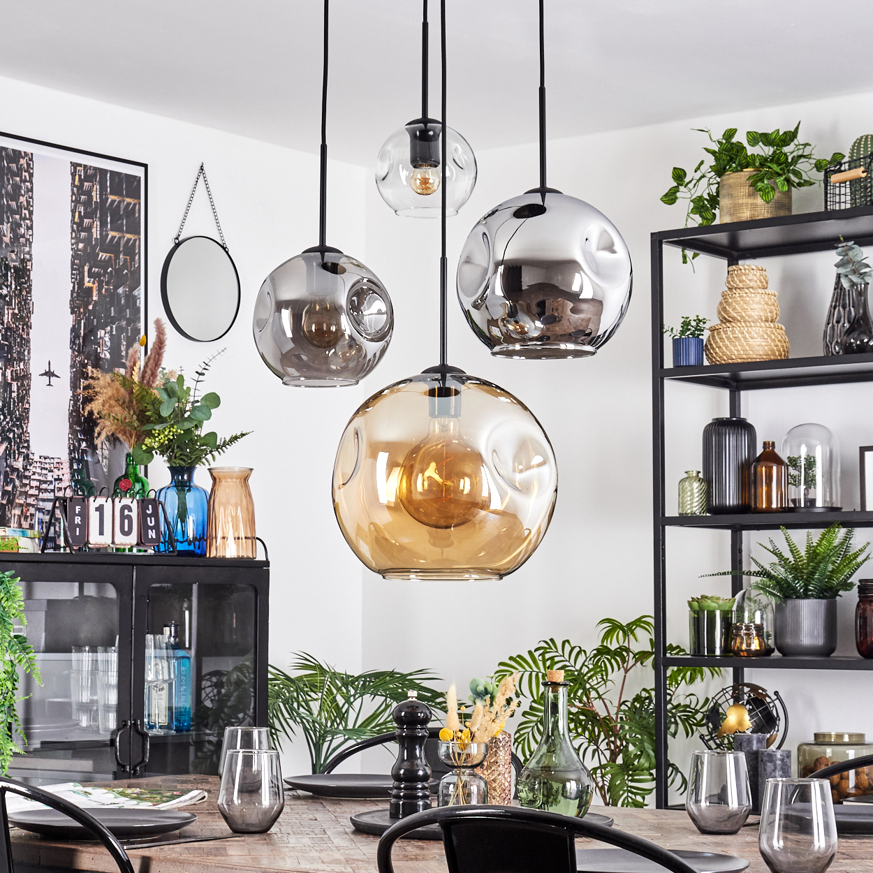 Esszimmer mit Pendelleuchten, gedecktem Tisch, Pflanzen und einem schwarzen Metallregal mit Dekorationen.