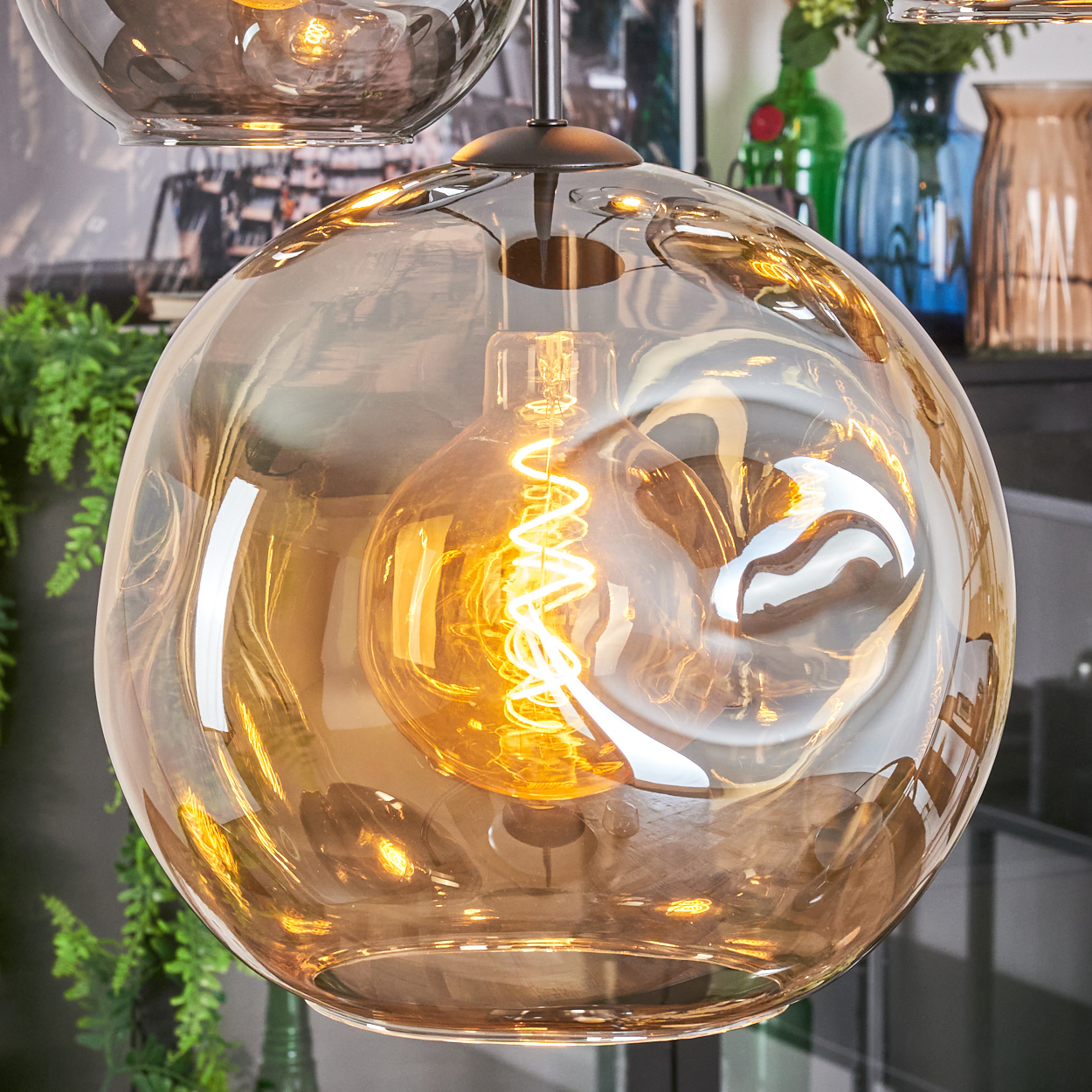 Nahaufnahme einer Glas-Hängelampe mit einer leuchtenden, spiralförmigen Glühbirne. Die Lampe hat einen bräunlichen Farbton. Vasen und Pflanzen im Hintergrund.