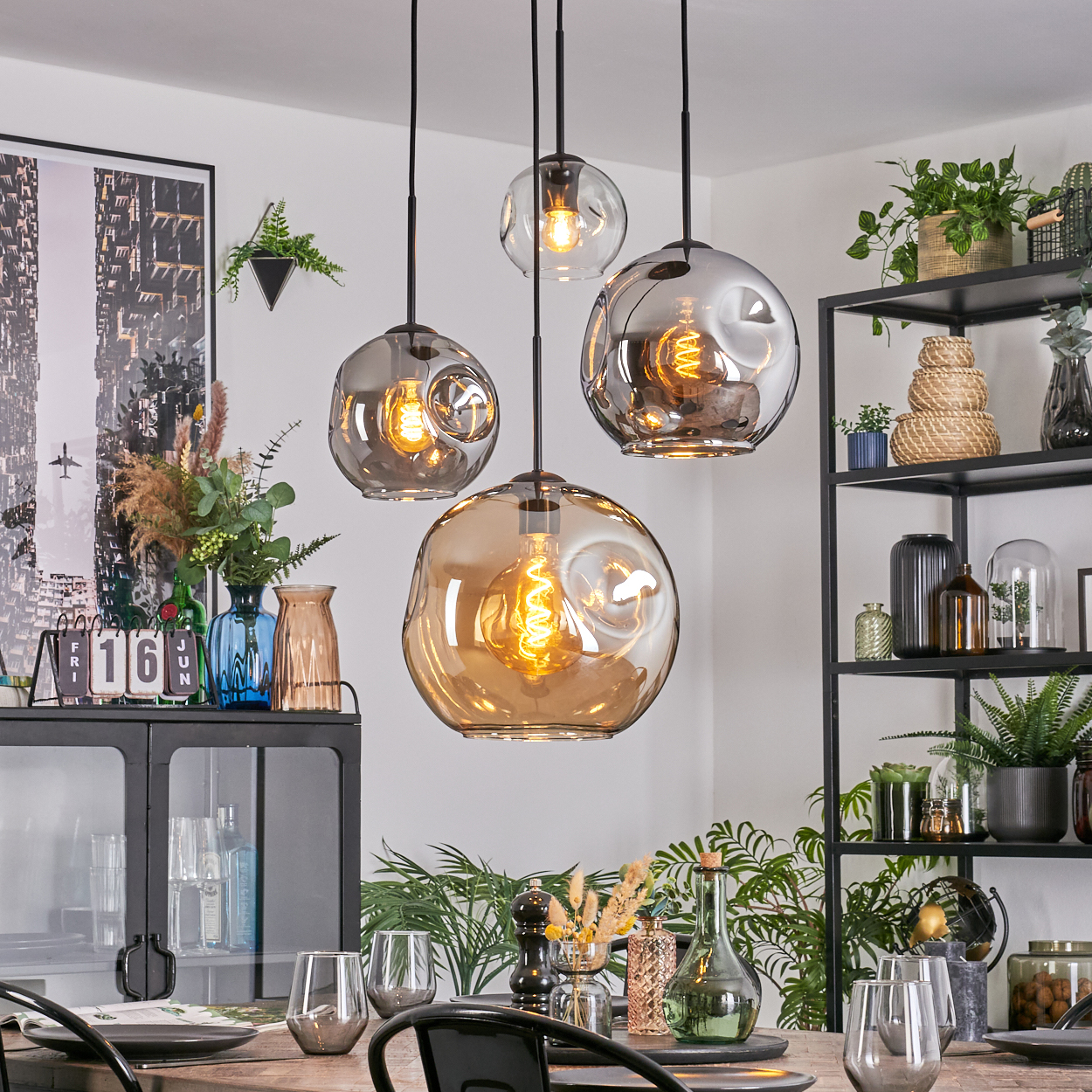 Esszimmer mit vier Glaskronleuchtern. Ein Tisch ist mit Gläsern und Tellern gedeckt. Regale und Pflanzen sind im Hintergrund.