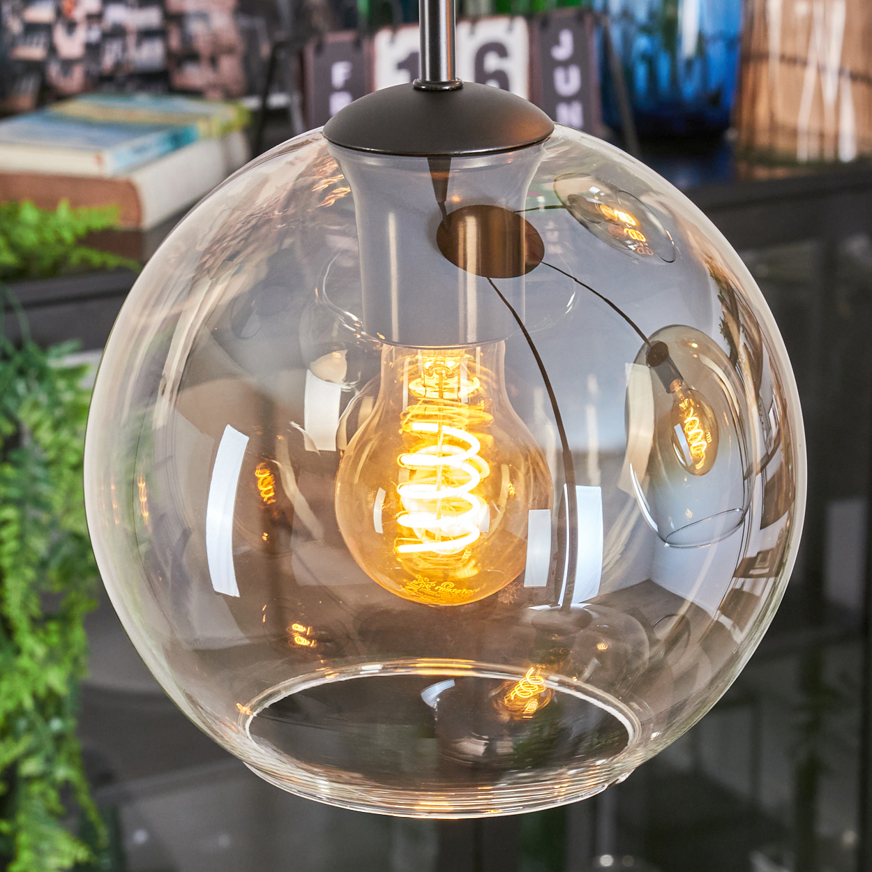 Eine klare, runde Glaslampe mit einer leuchtenden Wendelbirne. Die Lampe hängt an einer schwarzen Halterung, mit verschwommenen Hintergründen.
