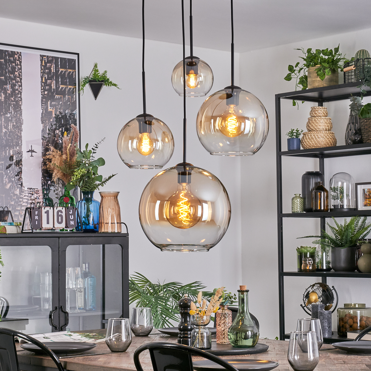 Esszimmer mit hängenden Kugelleuchten, Tisch mit Gläsern und ein schwarzes Metallregal mit Pflanzen und Dekor.