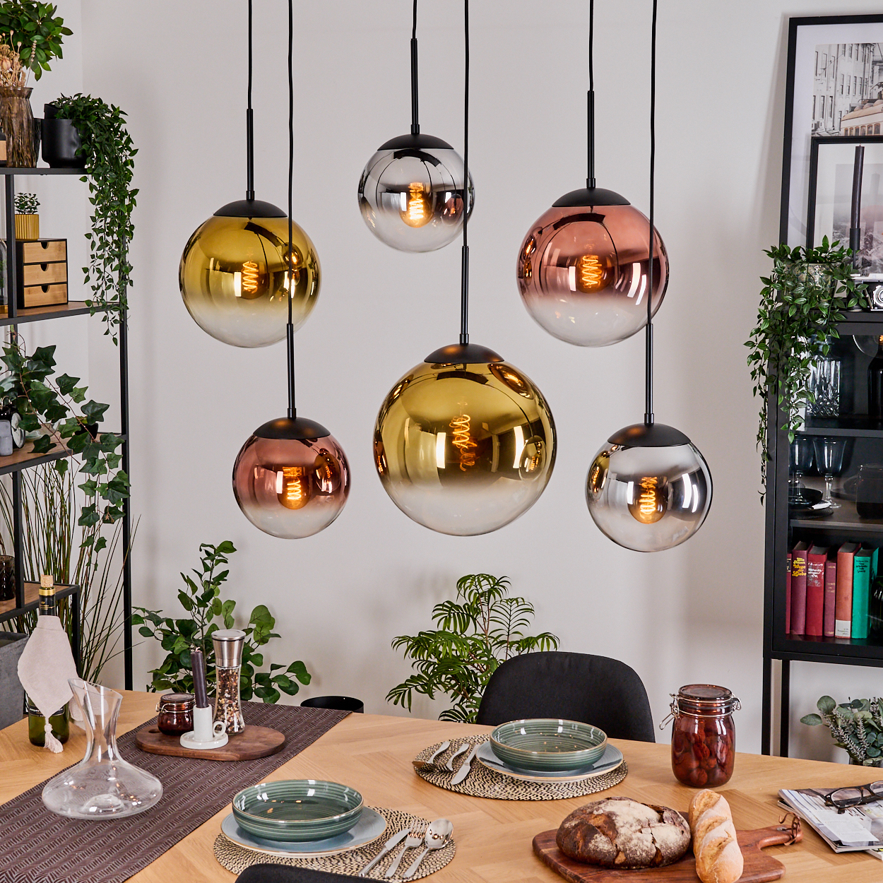 Esszimmer mit hängenden Kugelleuchten. Tisch mit Tellern, Brot und Glas. Pflanzen und Regale im Hintergrund.